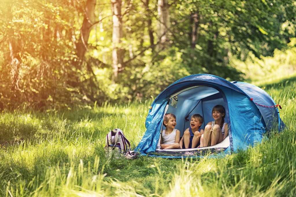 Little girl and brothers camping in a tent in a sunny forest